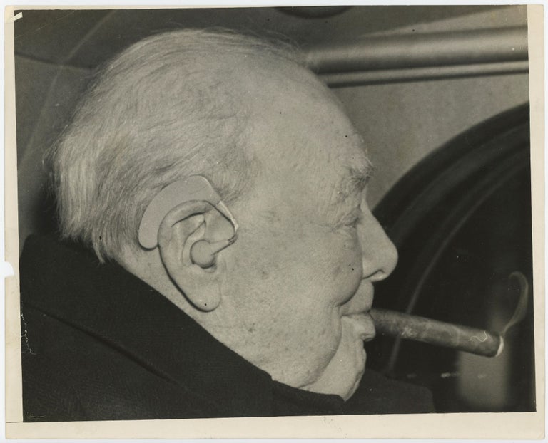 Item #005387 An original press photo of Sir Winston S. Churchill on 9 December 1958 leaving a lunch with Prime Minister Harold Macmillan, Churchill smoking a cigar, his hearing aid prominently visible.